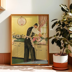 Könnte beinhalten: Ein gerahmter Druck im Vintage-Stil zeigt eine Person in einer Küche, die eine dunkle Weste und eine Schürze trägt und Geschirr spült. Ein kreisförmiger Wandbehang darüber zeigt den Text: He's a fucking battered wife! Look at him!