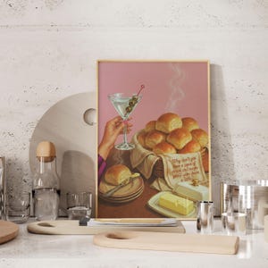 Pode incluir: Uma impressão emoldurada em estilo vintage com fundo rosa. Retrata um martini com azeitonas, uma cesta de pãezinhos, manteiga e um prato com um pãozinho. O texto na impressão diz: "Por que você não come um pedaço de pão e talvez se acalme um pouco?"
