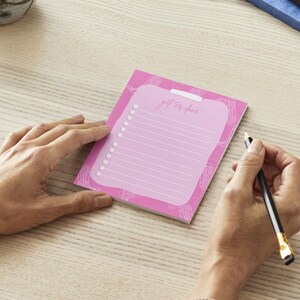 K&ouml;nnte beinhalten: Ein rosa Notizblock mit den Worten "get it done" und einem To-Do-Listen-Layout. Der Notizblock liegt auf einer hellen Holzoberfl&auml;che, mit einer Hand, die einen Bleistift h&auml;lt, um auf der Liste zu schreiben.