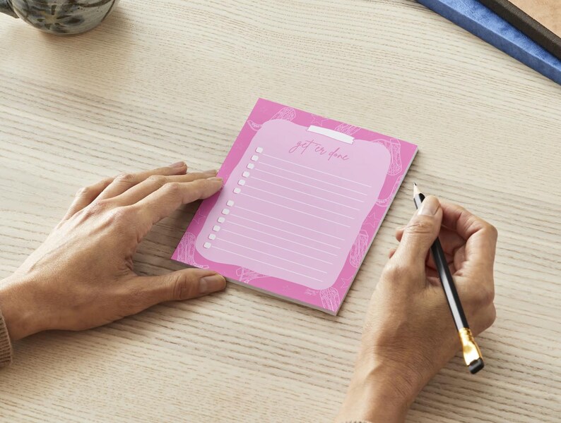 K&ouml;nnte beinhalten: Ein rosa Notizblock mit den Worten "get it done" und einem To-Do-Listen-Layout. Der Notizblock liegt auf einer hellen Holzoberfl&auml;che, mit einer Hand, die einen Bleistift h&auml;lt, um auf der Liste zu schreiben.