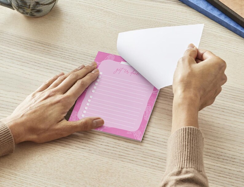 K&ouml;nnte beinhalten: Ein rosa Notizblock mit linierten Seiten und einem floralen Design. Das oberste Blatt wird abgezogen und zeigt den Text "get er done". Der Notizblock liegt auf einer hellen Holzoberfl&auml;che.