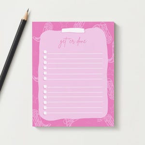 K&ouml;nnte beinhalten: Ein rosa Notizblock mit einer hellrosa To-Do-Liste und dem handschriftlichen Schriftzug &bdquo;get 'er done&ldquo;. Der Notizblock hat ein Cowboy-Stiefel- und Sternmuster. Ein schwarzer Bleistift mit goldener Zwinge liegt auf der wei&szlig;en Oberfl&auml;che.