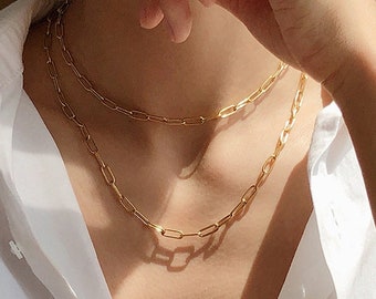 May include: A gold chain necklace with two layers of linked chains. The necklace is worn by a person with a white shirt.