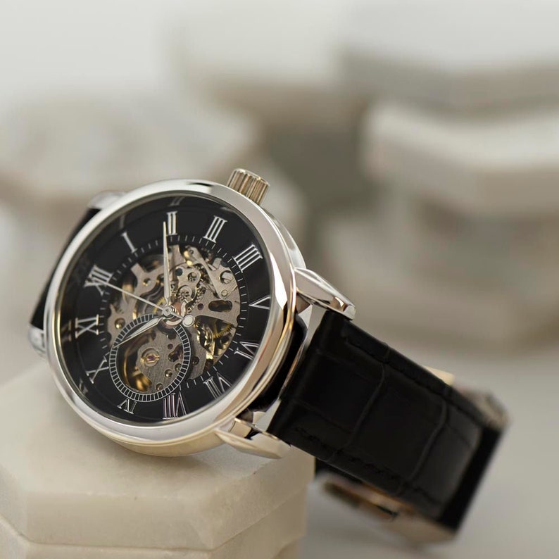 May include: A silver-toned watch with a black face and Roman numeral hour markers. The watch has a skeletonized movement and a black leather band.
