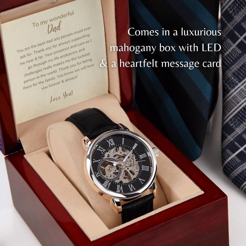 May include: A black leather wristwatch with a silver and gold face and Roman numeral markings. The watch is in a mahogany box with a heartfelt message card. The card reads "To my wonderful Dad".