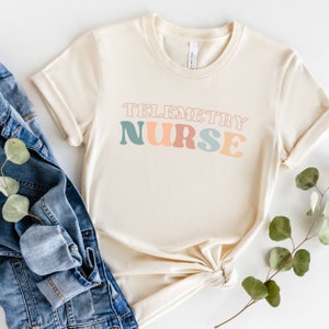 Könnte beinhalten: Ein beiges T-Shirt mit dem Text "Telemetry Nurse" in bunten Retro-Lettern.