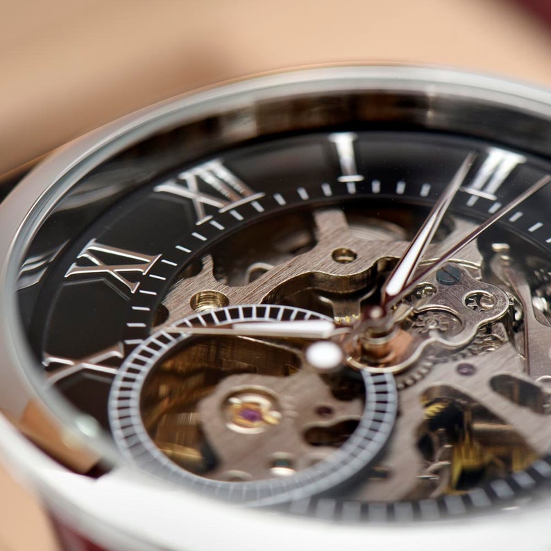 May include: A silver watch with a black face and Roman numeral hour markers. The watch has a skeletonized design, revealing the intricate inner workings of the movement.