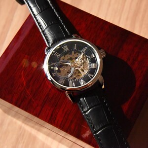 May include: A silver and black skeleton watch with Roman numeral markings on a black leather band. The watch is in a red wooden box.