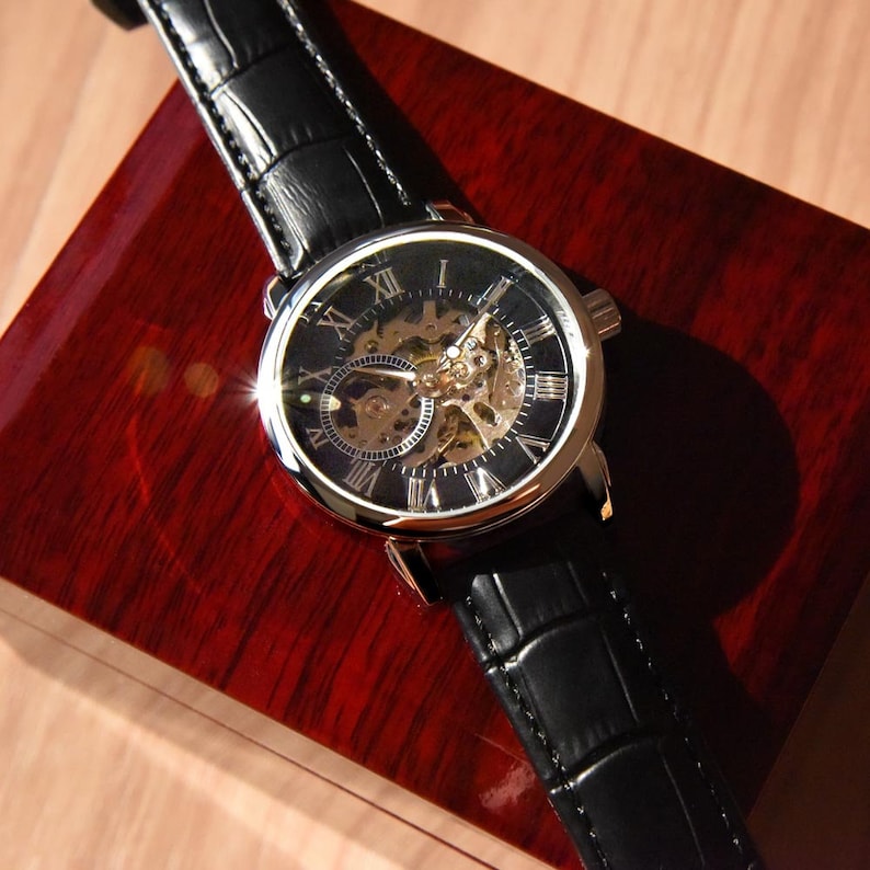 May include: A silver and black skeleton watch with Roman numeral markings on a black leather band. The watch is in a red wooden box.