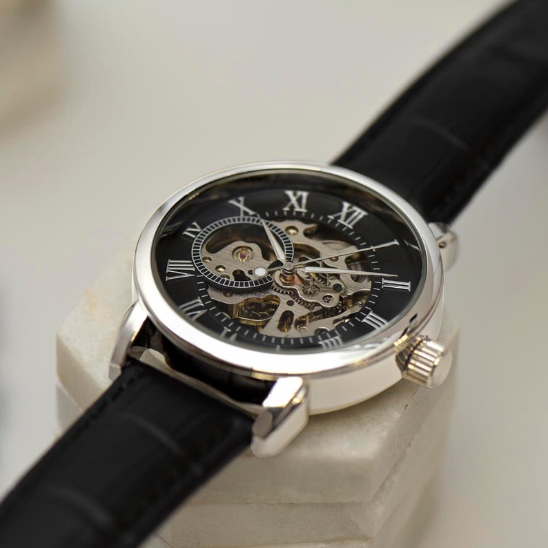 May include: A silver and black skeleton watch with Roman numeral markings on the face and a black leather band.