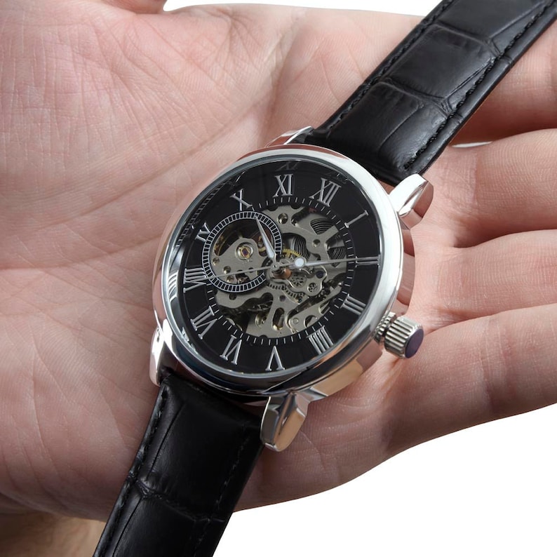 May include: A silver-toned wristwatch with a black face and Roman numeral hour markers. The watch has a skeletonized design, revealing the inner workings of the movement. The watch is on a black leather band.