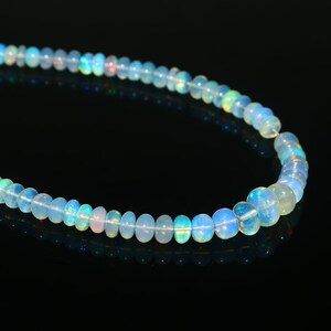 May include: A strand of opalescent opal beads. The beads are round and vary in size, strung together on a delicate thread. The opals display a range of colors, including blue, green, and flashes of iridescent hues. The background is black.