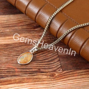 May include: A silver necklace with a teardrop-shaped pendant. The pendant has an opal stone with orange, yellow, and blue hues. The necklace is displayed on a wooden surface with the text "GemsHeavenIn".