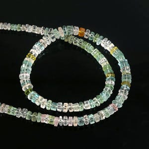 Könnte beinhalten: Eine Halskette aus kleinen, quadratisch geschliffenen Edelsteinen in den Farbtönen Klar, Hellblau, Grün, Gelb und Rosa. Die Perlen sind aufgefädelt und bilden ein zartes und farbenfrohes Accessoire. Der Hintergrund ist schwarz.