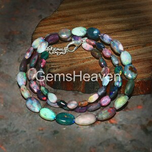 Peut inclure: Un collier d'opales multicolores avec un fermoir argenté. Les opales de forme ovale présentent une variété de couleurs, notamment le bleu, le vert, le violet et le rose. Le collier est présenté sur une surface en bois, et le texte "GemsHeaven" est visible.