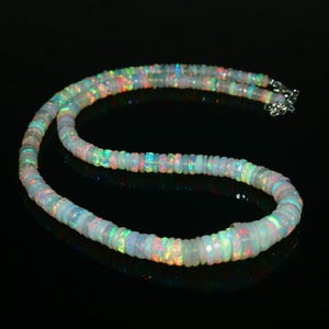 May include: A necklace composed of numerous small, iridescent opal beads. The beads display a range of colors, including blue, green, orange, and yellow, and are strung together with a silver clasp. The necklace is displayed against a black background.