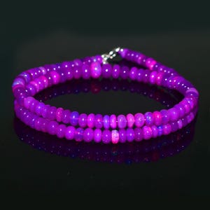 May include: A double-strand bracelet made of small, round, purple beads. The beads are polished and strung together with a silver clasp. The bracelet is shown on a reflective black surface.
