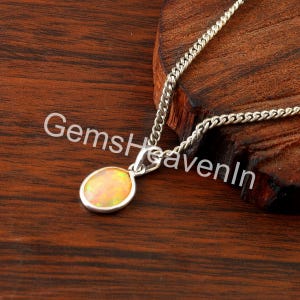 May include: A silver necklace with an oval opal pendant. The opal has yellow and orange tones. The chain is a curb style, and the pendant is set in a silver bezel. The background is a dark wood surface. The text "GemsHeavenIn" is visible.