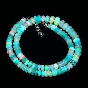 May include: A necklace composed of small, faceted opal beads in shades of blue, green, and pink. The beads are strung together with a silver-colored chain and clasp. The necklace is displayed against a black background.