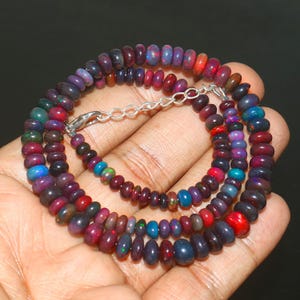 May include: A multi-strand necklace composed of small, polished beads in shades of purple, red, blue, and green. The beads are strung together with a silver-colored clasp. The necklace is displayed against a dark background.