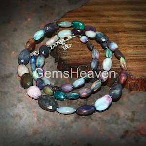 May include: A multi-colored opal necklace with oval-shaped beads. The necklace features a variety of colors, including blue, green, purple, and brown. The necklace is displayed on a wooden surface. The text "GemsHeaven" is visible.