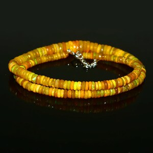 May include: A necklace composed of small, faceted, yellow and orange opal beads. The beads are strung together with a silver-colored clasp. The necklace is displayed on a black surface, with a slight reflection.