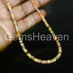 May include: A delicate necklace composed of small, colorful opal beads. The beads display a range of hues, including orange, yellow, green, and white. The necklace is held against a dark background, with the text "GemsHeaven" visible.