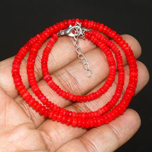 May include: A vibrant red beaded necklace with a silver-colored clasp and extender chain. The necklace is composed of small, round beads, creating a layered effect. The beads are a bright, solid red color.