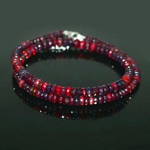 May include: A double-strand necklace composed of small, faceted beads in shades of red and purple. The beads are strung together, forming a circular shape. The necklace has a silver clasp. The background is black.