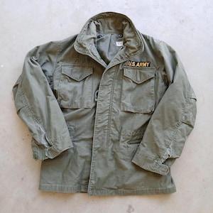 Può includere: Giacca militare verde oliva con quattro tasche frontali e collo. La giacca presenta una toppa "U.S. ARMY" sul petto destro. La giacca è realizzata in un tessuto resistente e ha una vita regolabile.