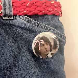 May include: Close-up of a pair of blue denim jeans with a red braided belt. A round button with a photo of a man with long hair and a pink bow is pinned to the jeans. The jeans are worn and have a vintage look.