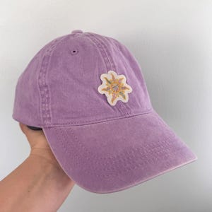 May include: A lavender baseball cap with a decorative patch. The patch is a starburst design with yellow, pink, and green accents. The cap is made of a soft, worn fabric.