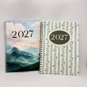 May include: Two spiral-bound planners for the year 2027. The left planner features a mountain landscape with a cloudy sky. The right planner has a green and white vertical striped pattern with floral accents and a central oval label.