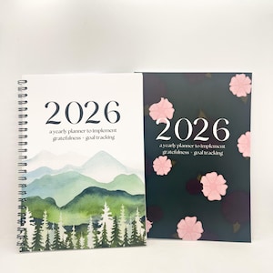 Può includere: Due agende annuali 2026. Una presenta un design di montagna e foresta ad acquerello, l'altra illustrazioni di fiori rosa su sfondo scuro. Entrambe le agende riportano il testo "a yearly planner to implement gratefulness + goal tracking."