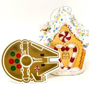 Può includere: Un ornamento di pan di zenzero dorato a forma di nave spaziale Millennium Falcon di Star Wars. L'ornamento è decorato con caramelle rosse e verdi.