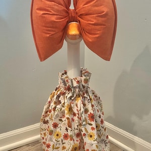 May include: A decorative goose dressed in a fall-themed outfit. It features a large orange bow, a white neck, and a skirt with a floral pattern of sunflowers, leaves, and acorns. The goose is standing on a wooden floor.