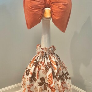 May include: A decorative goose statue wearing an orange bow and a dress with a bow pattern in shades of brown and orange. The goose has a white neck and a green base.