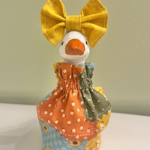May include: A white goose figurine wearing a bright yellow fabric bow on its head and a gathered, patchwork-style fabric dress featuring orange, blue, and green floral and sunflower patterns with a ruffled neckline around the neck of the goose.