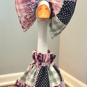 May include: A decorative goose figurine wearing a patchwork bow and dress. The bow and dress feature a mix of floral, polka dot, and gingham patterns in pink, navy, and green. The goose has an orange beak.