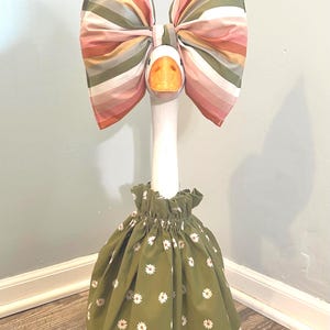May include: A decorative goose figurine with a large striped bow in shades of pink, orange, and green. The goose wears a green dress adorned with white daisy patterns. The figurine is positioned in a corner against a light blue wall.
