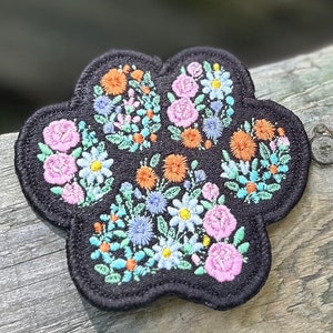 May include: A black paw print patch with embroidered colorful flowers. The patch is perfect for adding a touch of whimsy to clothing, bags, or other accessories.