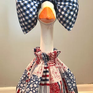 May include: A decorative goose figurine wearing a large blue and white gingham bow and a patchwork dress in red, white, and blue. The dress features floral, paisley, and American flag patterns.