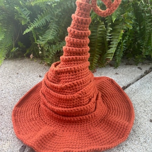 May include: A crocheted witch hat in a burnt orange color. The hat has a wide brim and a tall, pointed crown that curls at the top.