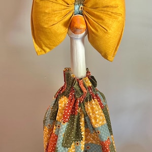 May include: A decorative goose figurine with a large yellow bow and a patchwork skirt. The goose has a white neck, an orange beak, and a colorful skirt with floral and geometric patterns. The bow is made of a textured yellow fabric.