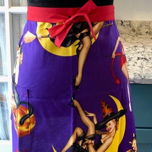 May include: A purple apron with a red waistband and bow. The apron features a pin-up style design with witches on a yellow crescent moon. The text "Style A" is visible in the upper right corner. The apron is displayed on a black mannequin.