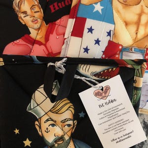 May include: Black pot holder with a sailor illustration, a football player, and a patriotic design. The pot holder has a loop for hanging and a tag that reads "Pot Holder" with care instructions. Made in the USA.