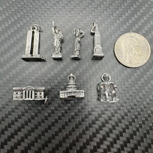 May include: A collection of silver-toned miniature charms, including the Twin Towers, the Statue of Liberty, the Washington Monument, the Empire State Building, the White House, the Capitol Building, and a seated figure. A quarter coin is also present.