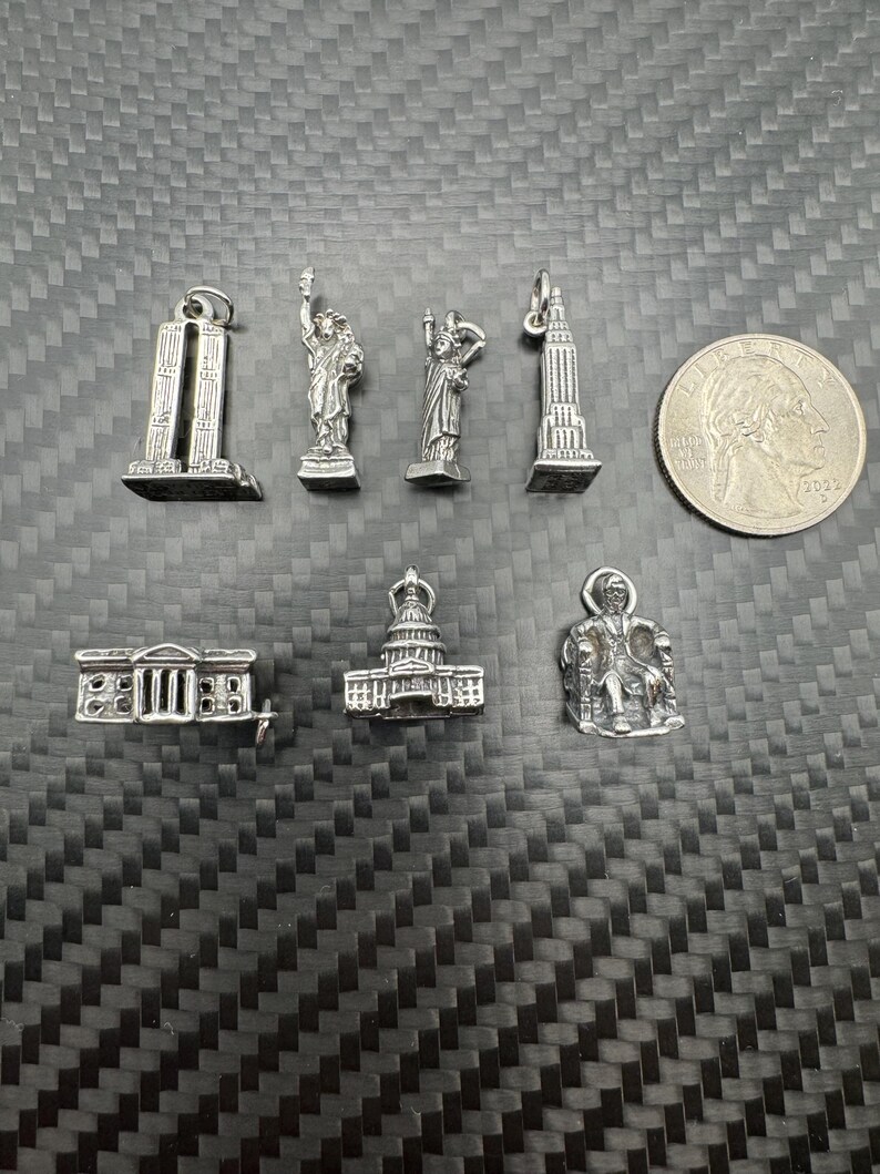 May include: A collection of silver-toned miniature charms, including the Twin Towers, the Statue of Liberty, the Washington Monument, the Empire State Building, the White House, the Capitol Building, and a seated figure. A quarter coin is also present.