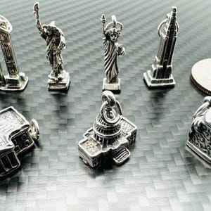 May include: A collection of silver-toned miniature charms depicting iconic American landmarks, including the Twin Towers, the Statue of Liberty, the Empire State Building, the Capitol Building, and a seated figure. A US quarter coin is also present.