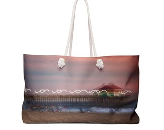 May include: A large white canvas tote bag with a pink and orange sunset scene printed on it. The scene features a pier with a building on it and the ocean in the background. The bag has white rope handles.